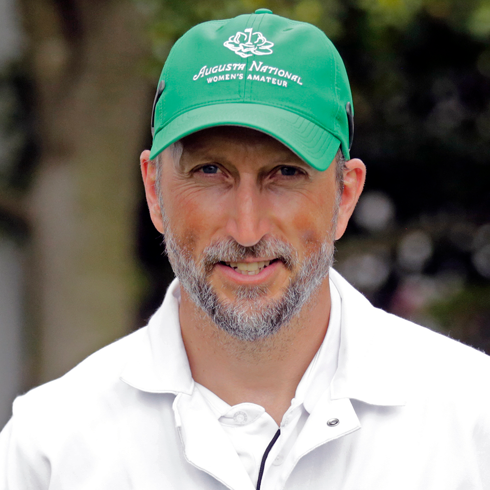 Man med skägg bär en grön keps med texten "Augusta National Women's Amateur" och vit jacka, utomhus med suddig bakgrund.