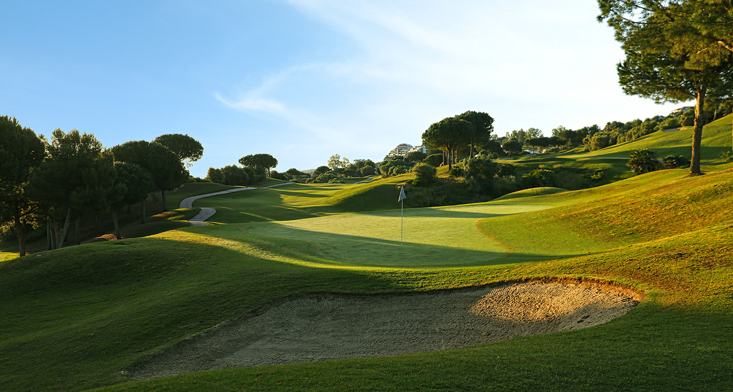 En golfbana med grönt gräs, sandbunker i förgrunden, flagga på green och träd runt banan under klar himmel.