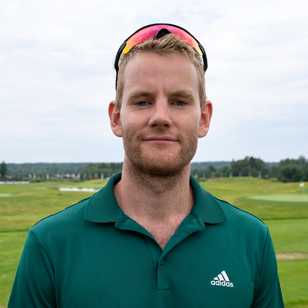 Man med kort blont hår och skägg bär grön Adidas-tröja och solglasögon på huvudet, står på en golfbana utomhus.