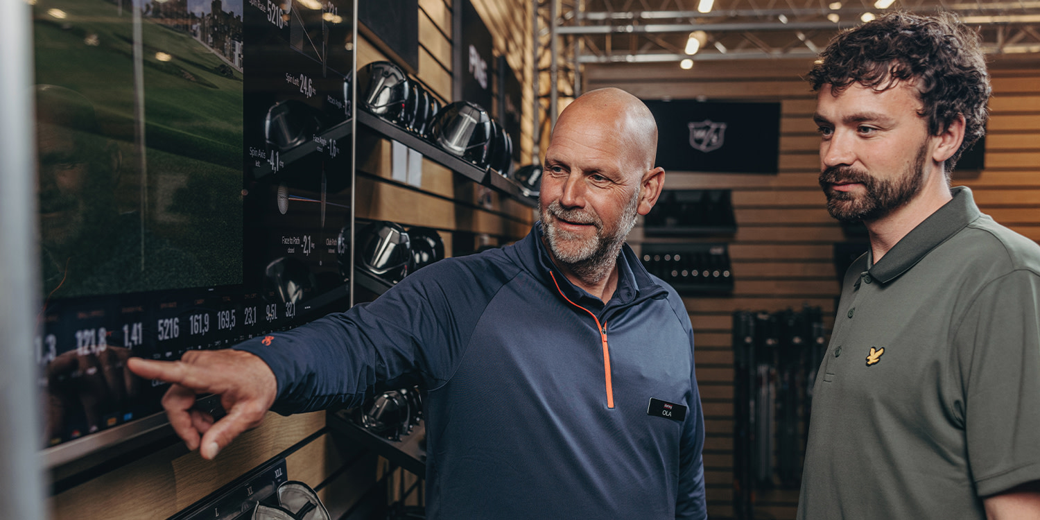 Two men are standing in a golf shop; one is pointing at a screen displaying golf data while the other looks on.