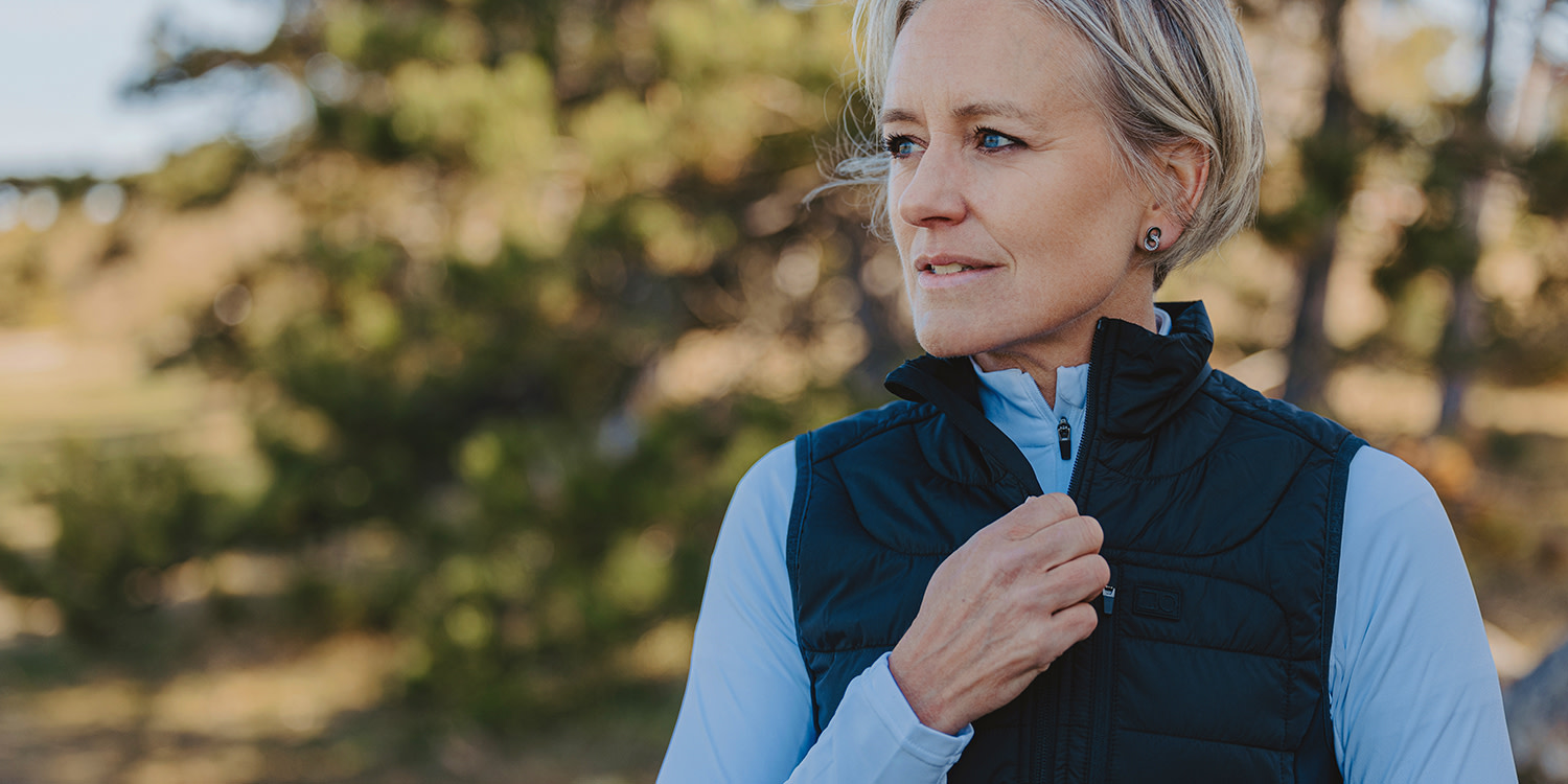 Kvinna med kort blont hår drar upp dragkedjan på sin svarta väst utomhus i en solig skogsmiljö.