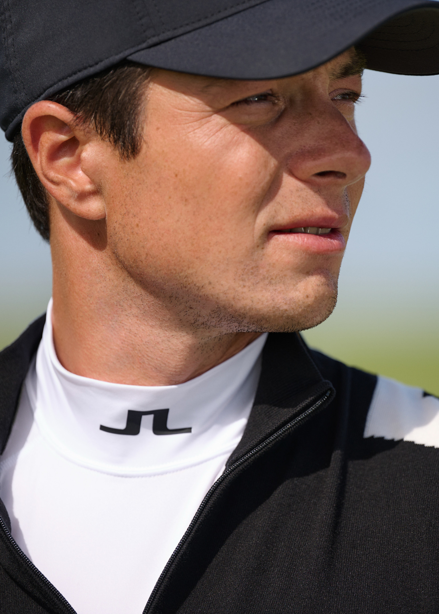 Man med svart keps och svartvit jacka tittar åt sidan utomhus, JL-logotyp syns på hans vita krage.