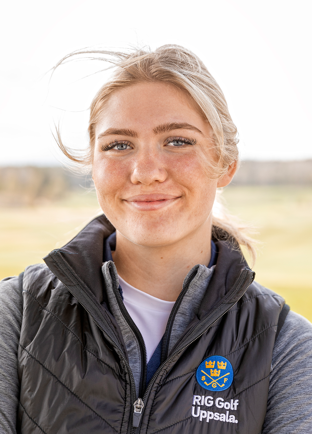 Ung kvinna med blont hår ler, bär svart väst med texten "RIG Golf Uppsala" och står utomhus på en golfbana.