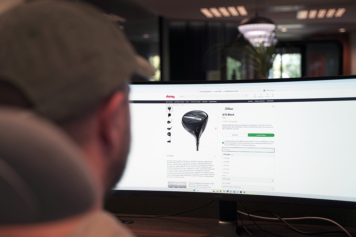 A person wearing a cap is looking at a computer screen and visiting Dormy's website to view a golf club, the GT2 Black.