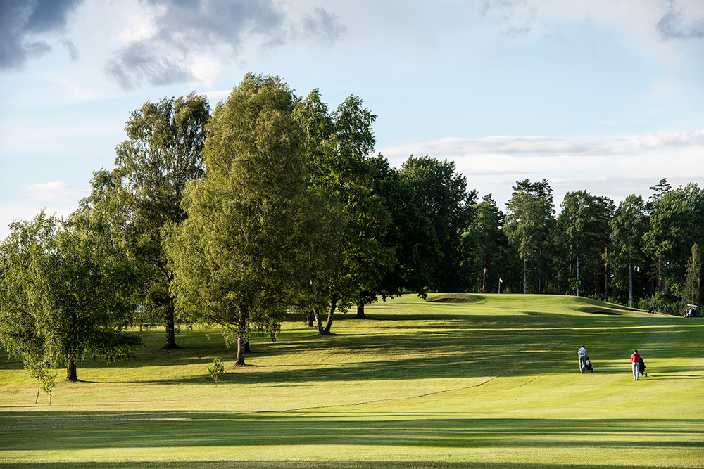Två personer går med golfbagar på en grön golfbana omgiven av träd under en delvis molnig himmel.