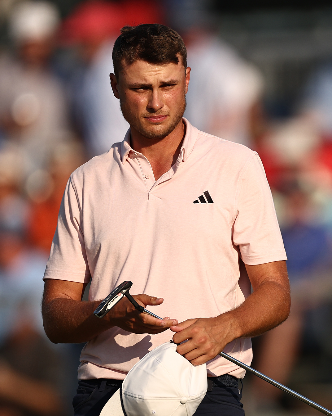 Man i ljusrosa pikétröja med Adidas-logga håller en golfklubba och vit keps, står utomhus på en golfbana.