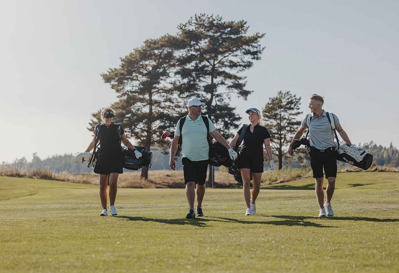 Fyra personer går tillsammans på en golfbana, bär golfbagar och ler mot varandra under en solig dag med träd i bakgrunden.