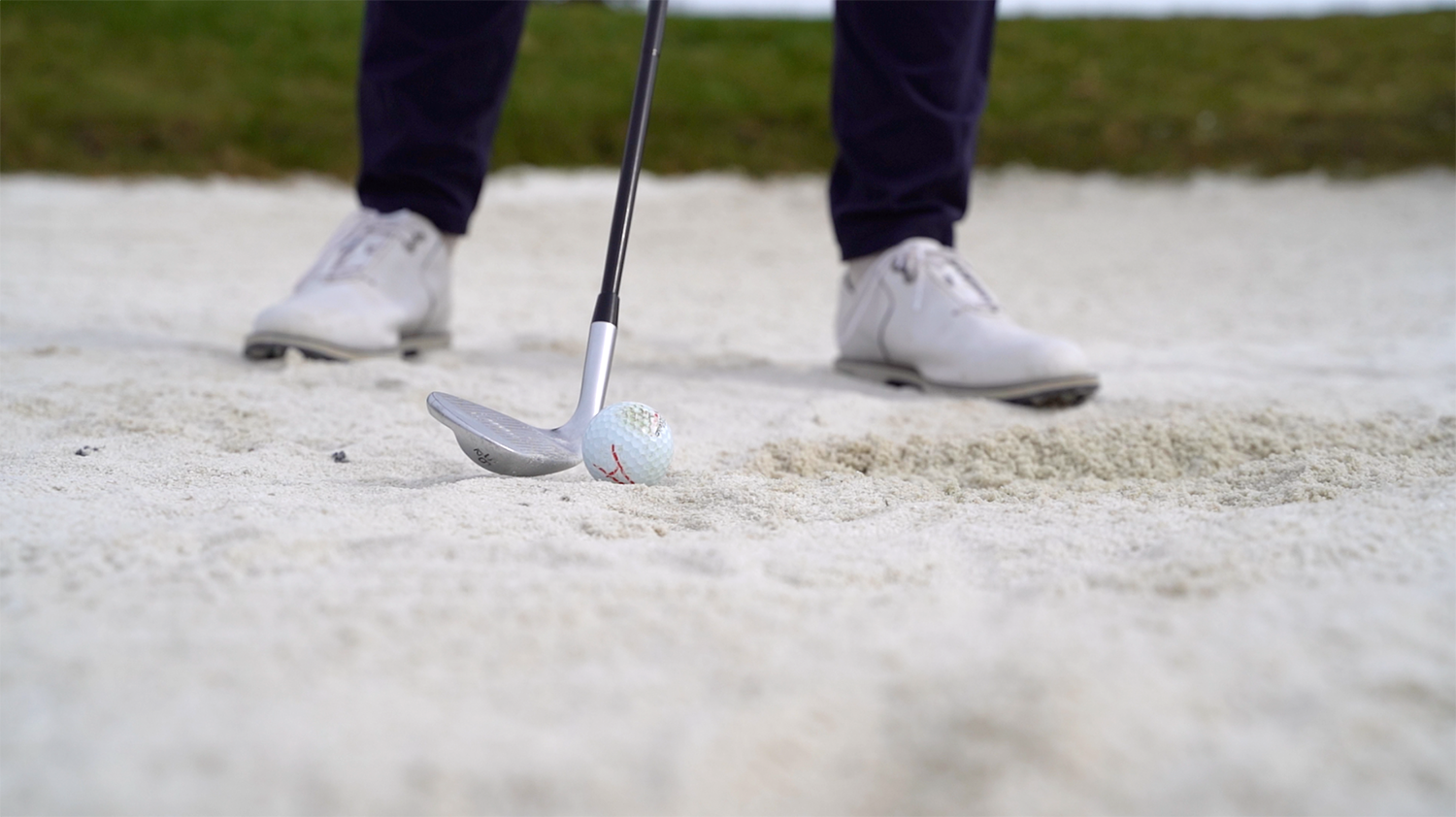 Golfklubba slår mot en golfboll i en sandbunker, med en persons fötter i vita skor i bakgrunden.