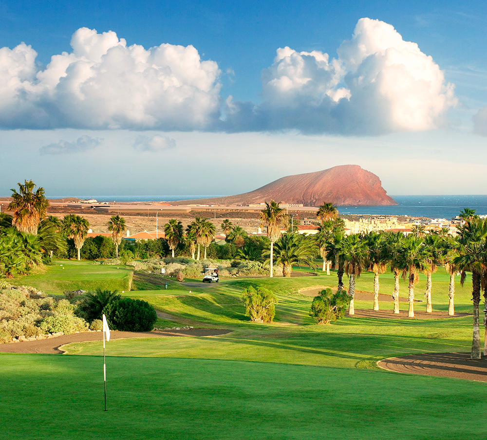 Golfbana med grönt gräs och palmer, golfbil i bakgrunden, hav och ett berg syns under blå himmel med moln.