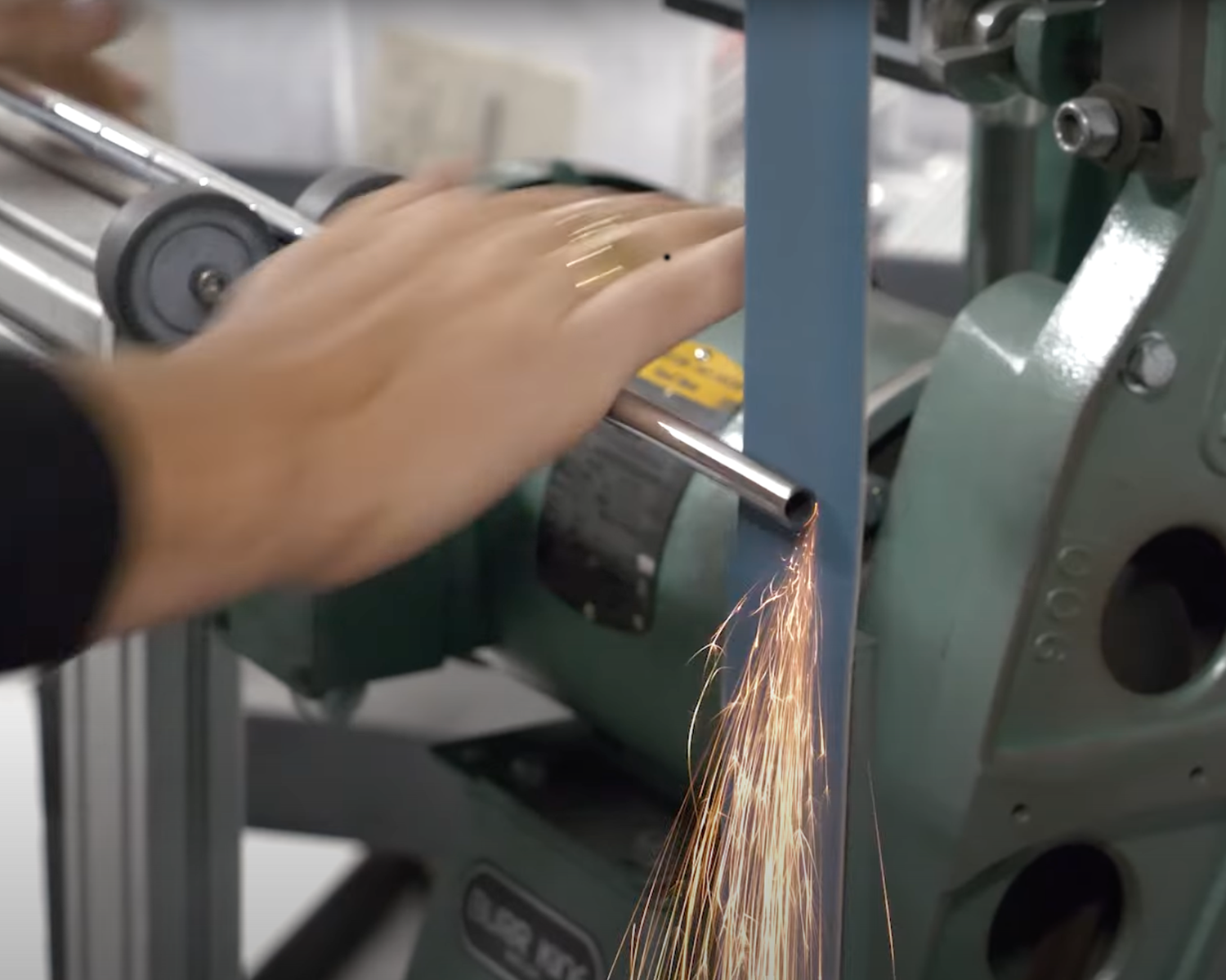 En hand håller ett metallrör mot ett roterande band, gnistor flyger när röret slipas i en verkstadsmiljö.