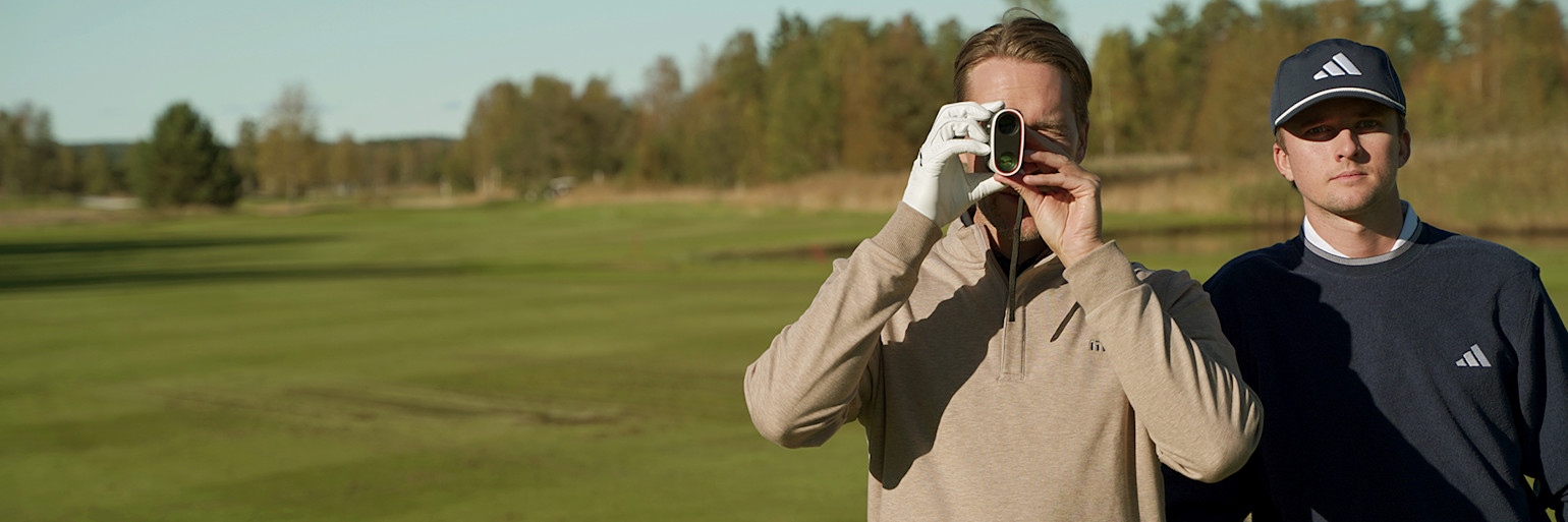 Two men are standing on a golf course; one is using a rangefinder while the other looks towards the camera, surrounded by greenery and trees.