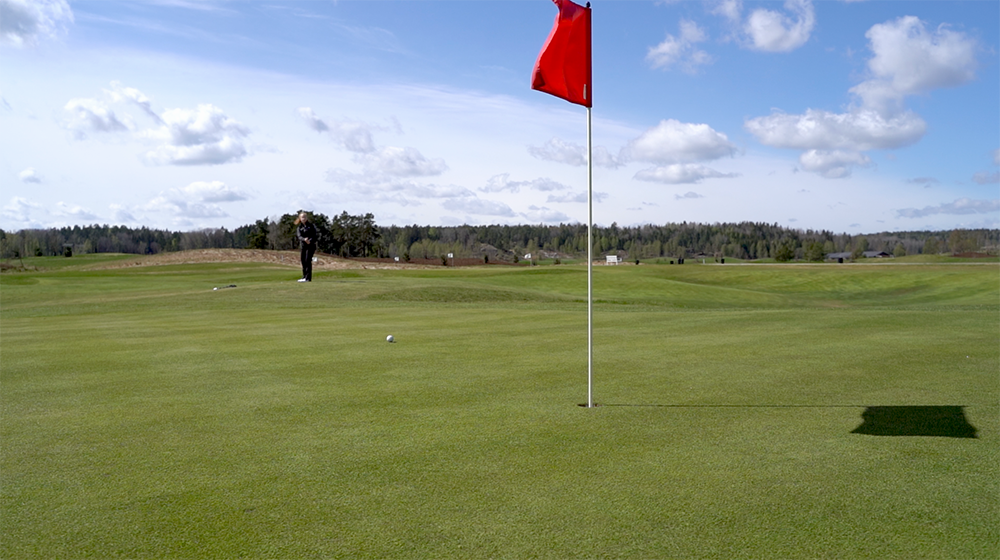 Röd flagga står i golfhål på green, golfboll ligger nära och en person förbereder slag på avstånd under blå himmel.