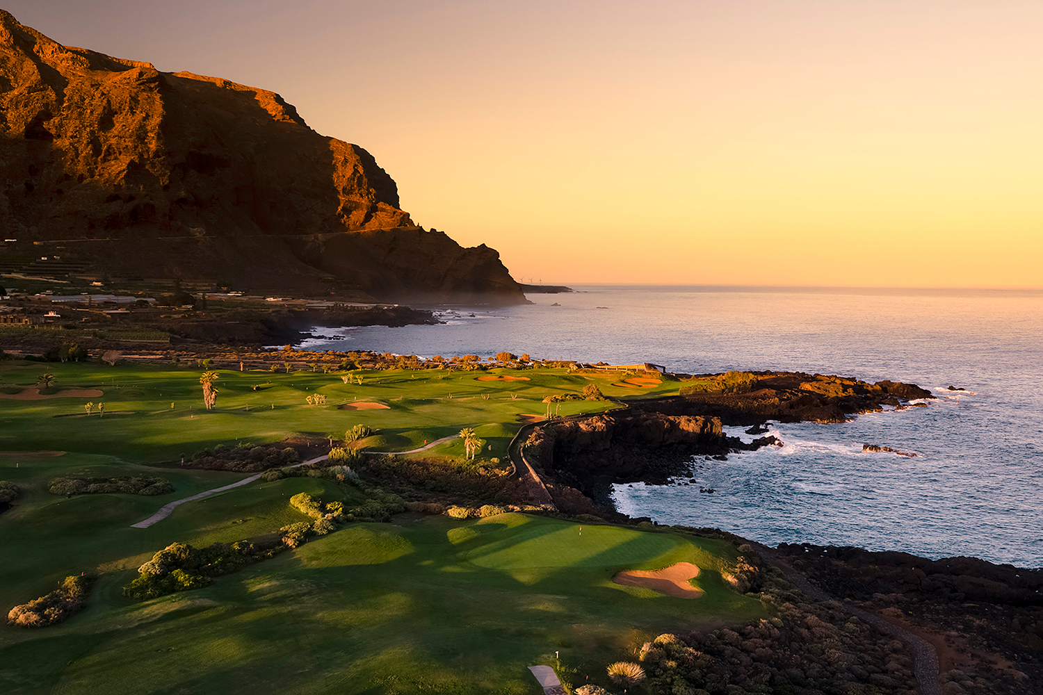 Golfbana med gröna fairways vid havet, omgiven av klippor och berg i solnedgångens varma ljus.