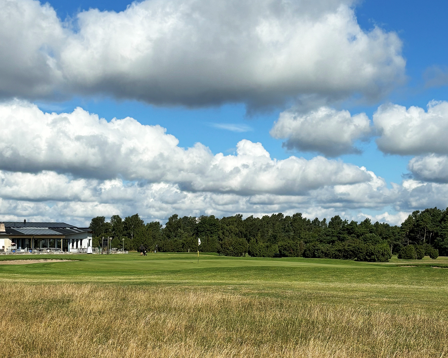 Golfbana med klubbhus till vänster, grönt gräs, flagga på green och träd i bakgrunden under molnig himmel.