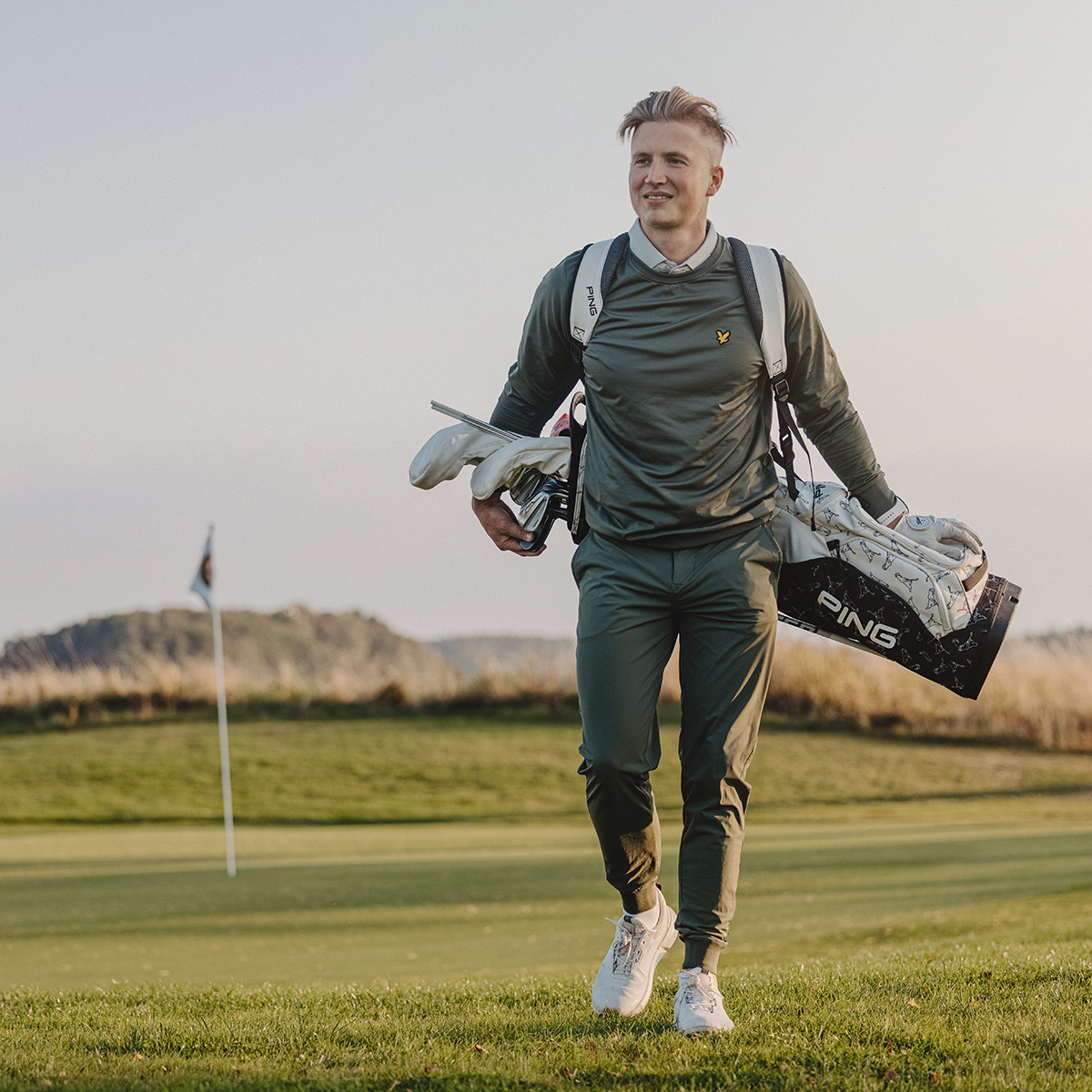 Man bär en golfbag med PING-logotyp och golfklubbor på en golfbana, med flagga och gräs i bakgrunden.