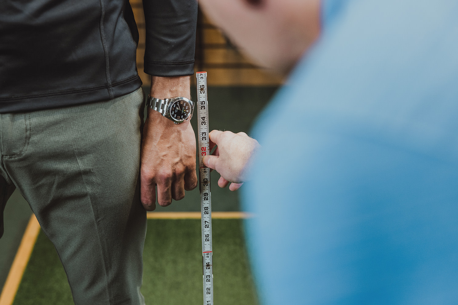 En person mäter en annan persons arm med ett måttband, där den ena bär en silverfärgad klocka och gröna byxor.