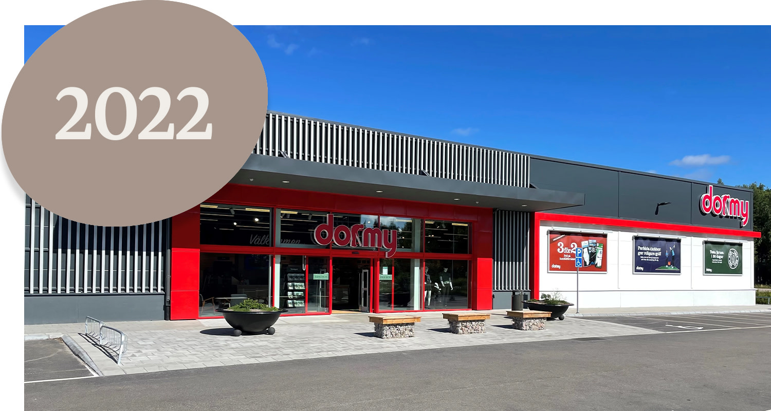 Shop with a red entrance and the sign "dormy" on the facade, blue sky, and a large beige circle with the text "2022" in the corner.