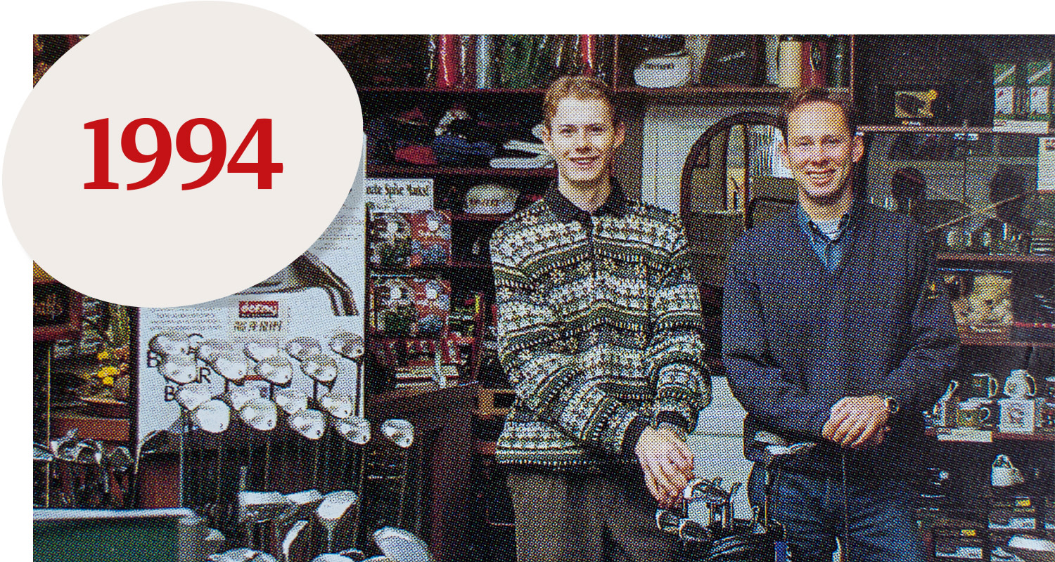 Two smiling men stand in a golf shop surrounded by golf clubs and accessories, with the text "1994" in red in one corner.