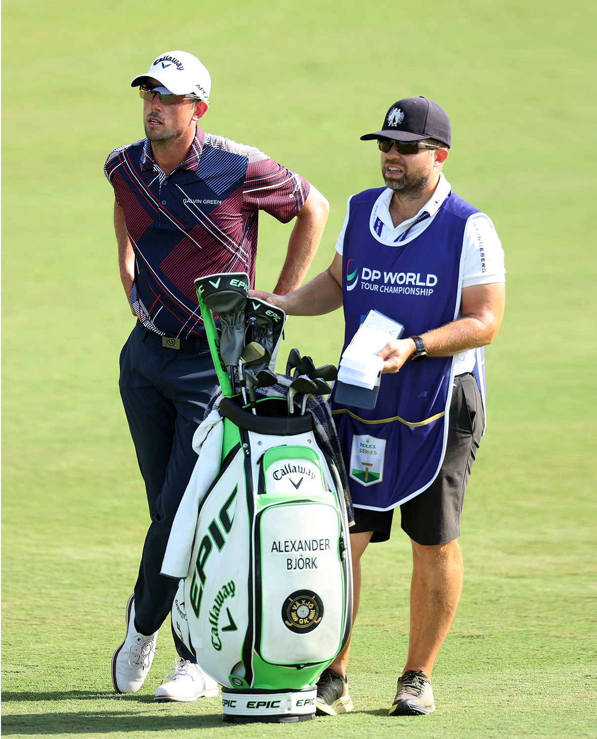 En golfspelare och en caddie står på en golfbana bredvid en golfbag märkt "Alexander Björk" och "Callaway EPIC".