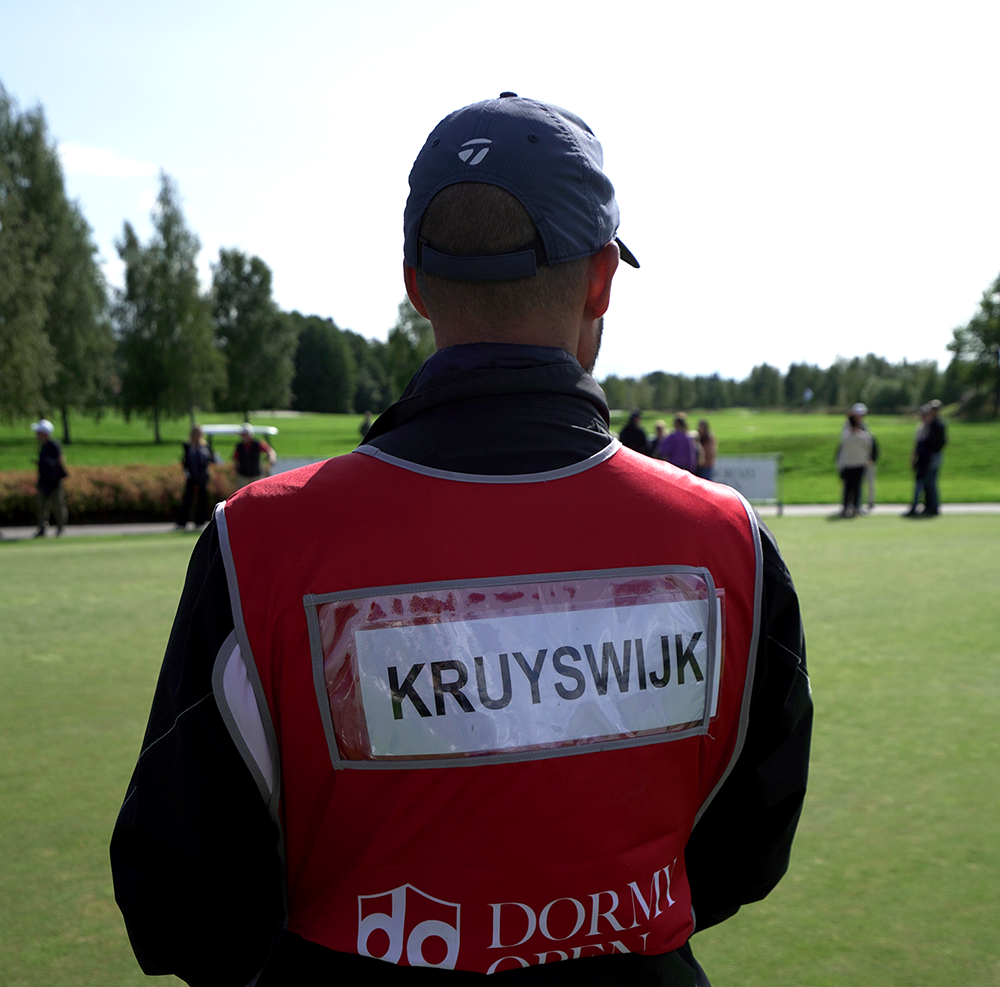En caddie med röd väst med texten "KRUYSWIJK" står på en golfbana med människor och träd i bakgrunden.