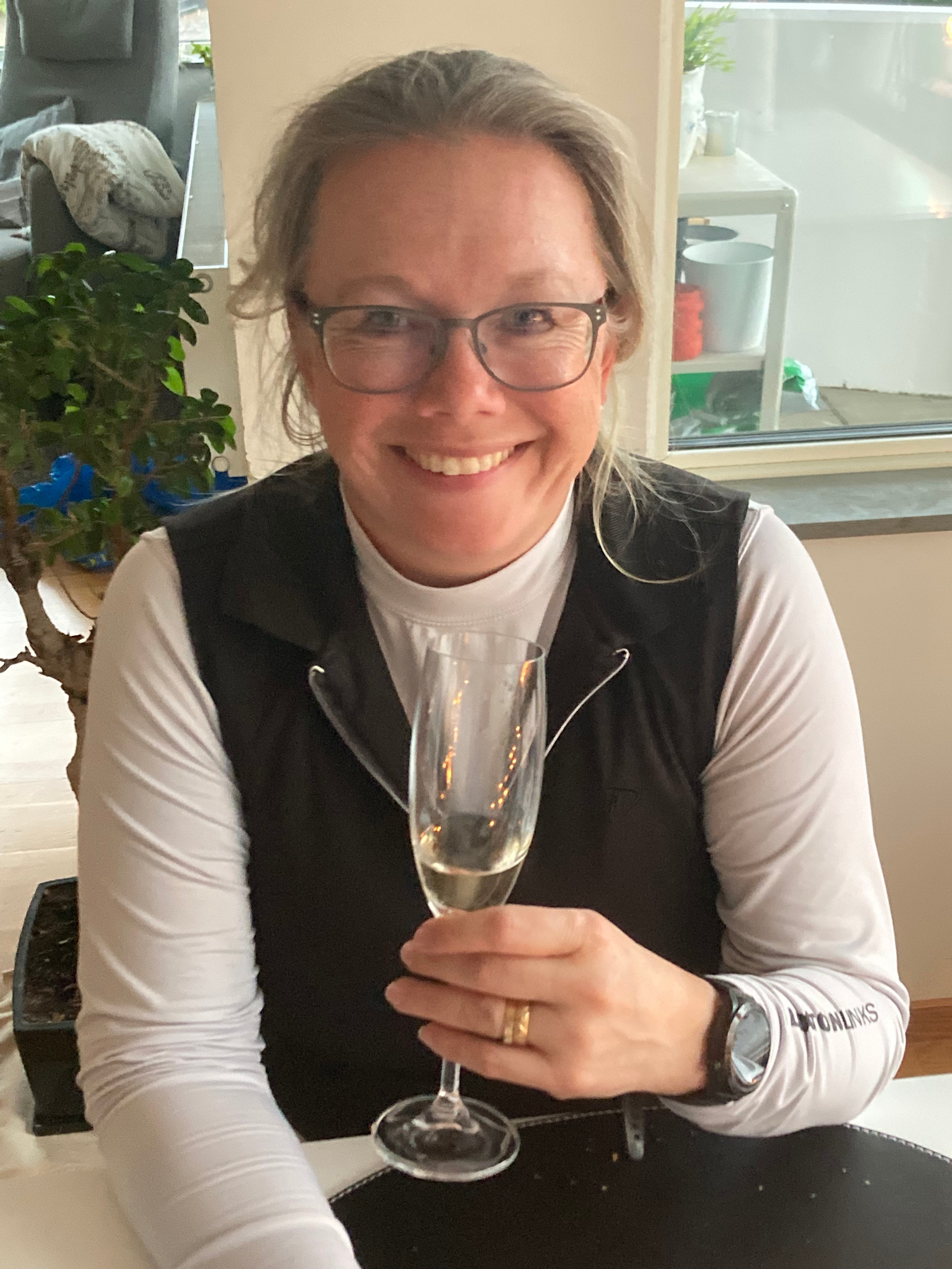 Leende kvinna med glasögon håller ett glas mousserande vin vid ett bord inomhus.