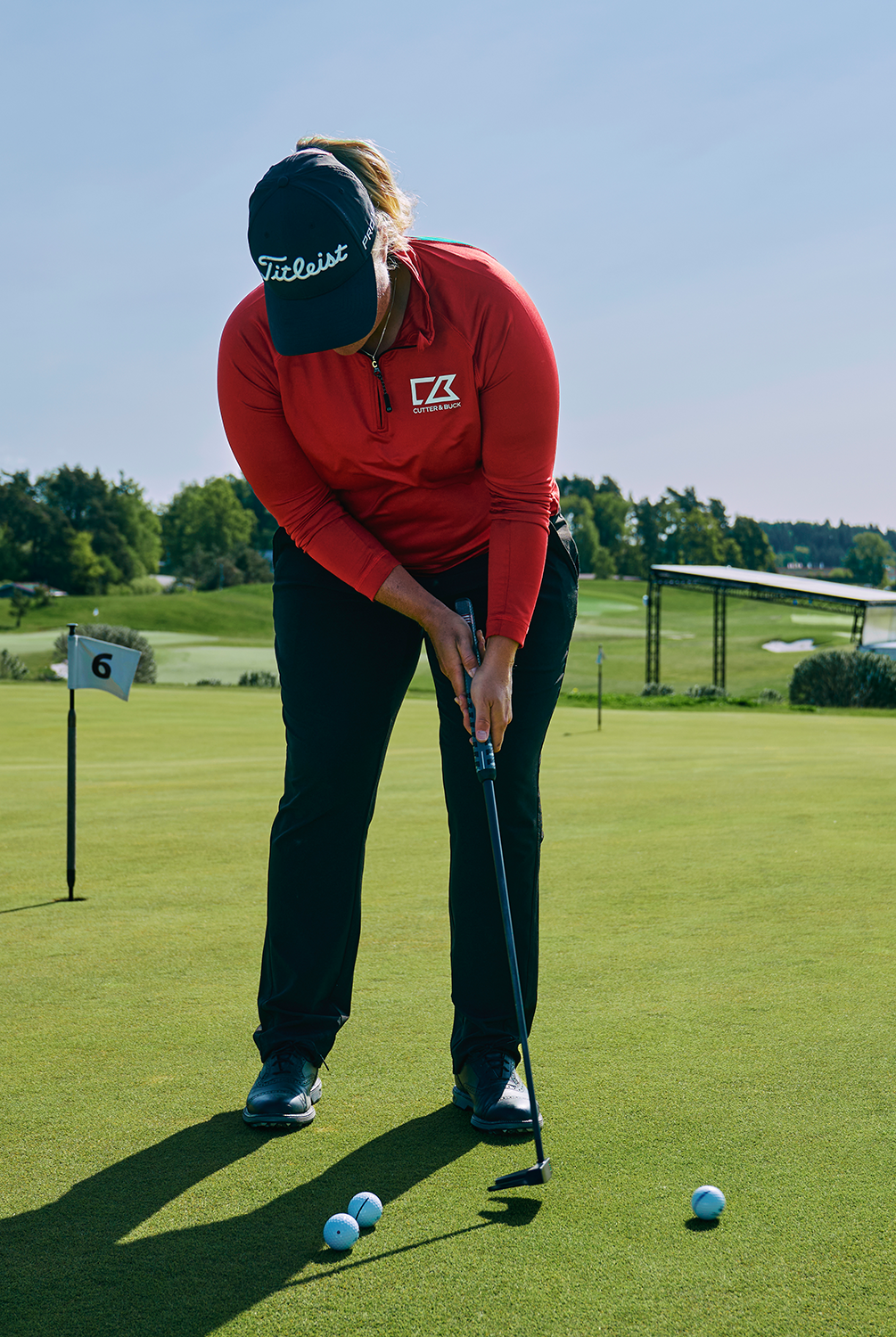 Person slår en putt på en golfgreen, iklädd röd tröja med Cutter & Buck-logga och mörk keps, tre golfbollar på marken.
