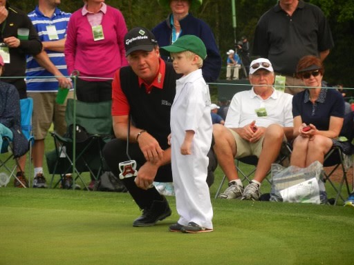 En man i golfkläder hukar sig bredvid ett litet barn i vit overall och grön keps på en golfbana, med åskådare i bakgrunden.