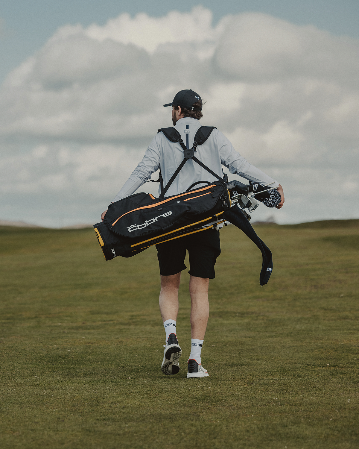 Man bär en Cobra-golfbag över axeln och går på en grön golfbana under en molnig himmel.