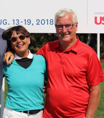 Två äldre personer står bredvid varandra utomhus och ler, med en skylt i bakgrunden som visar datumet 13-19 augusti 2018.