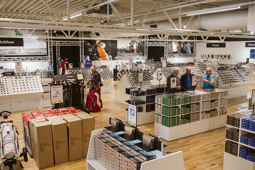 Flera personer tittar på golfklubbor och tillbehör i en välsorterad golfbutik med hyllor och stora reklamskyltar på väggarna.