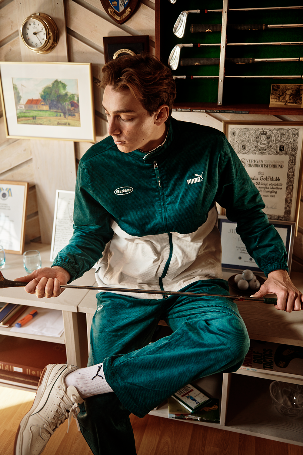 En person i grönvit Puma-träningsoverall sitter på en bänk och håller en golfklubba, omgiven av golfpriser och diplom på väggen.