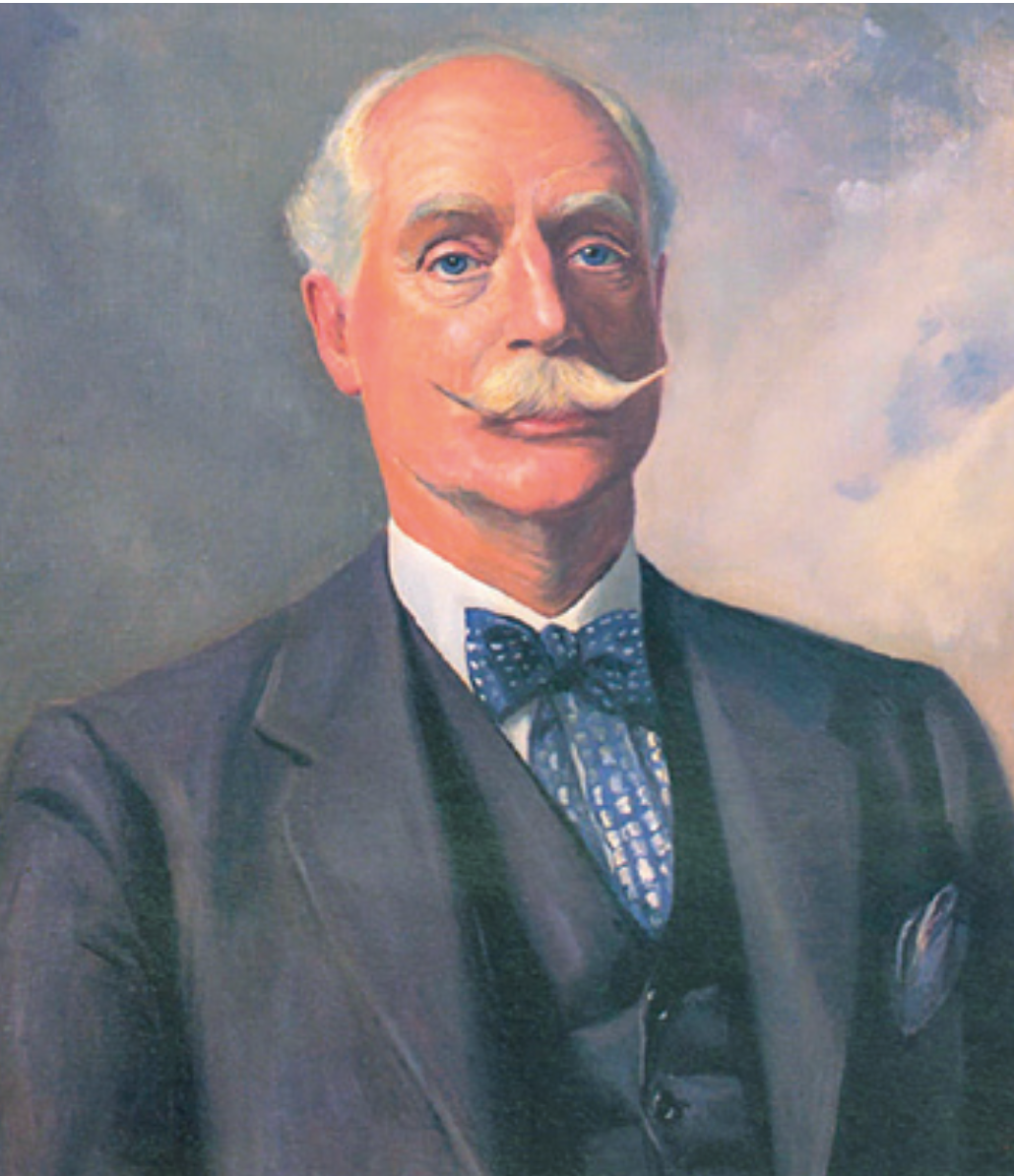 Äldre man med vitt hår och mustasch bär en mörk kostym, väst, vit skjorta och blå fluga mot en målad bakgrund.