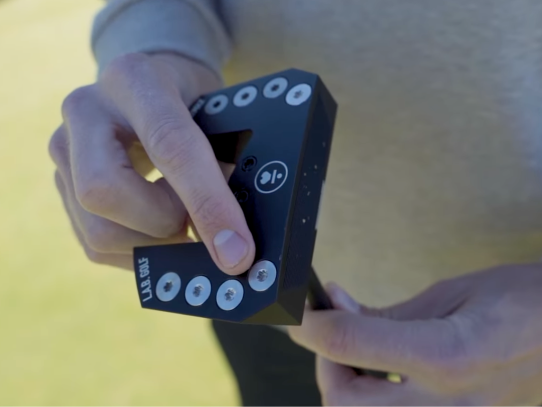 En person håller i ett svart golfputterhuvud med texten "L.A.B. GOLF" och flera silverfärgade vikter på klubbhuvudet.