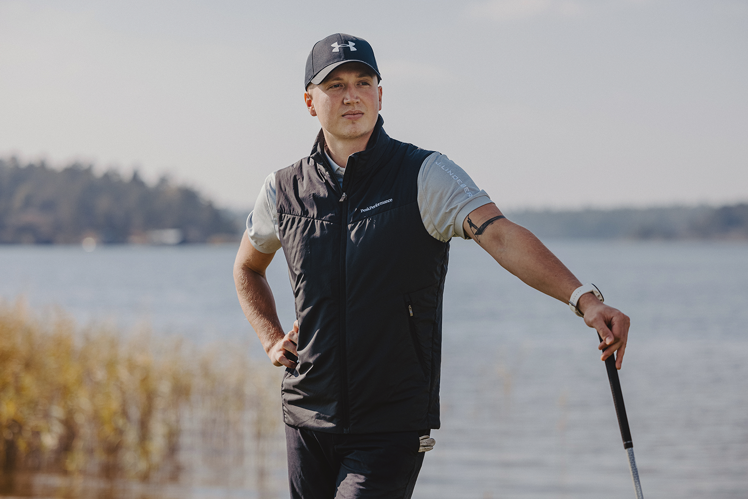 Man i golfkläder står avslappnat med en golfklubba vid en sjö, med natur och vatten i bakgrunden.