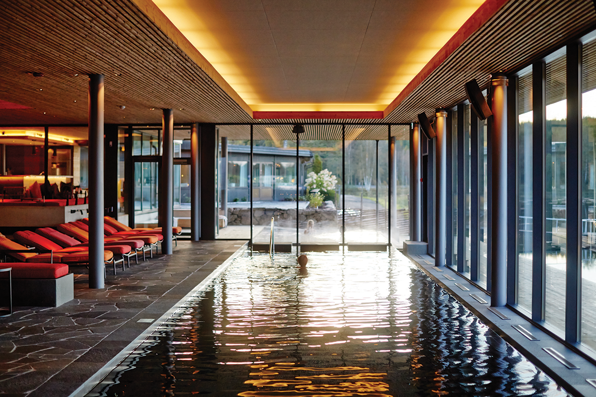 Person simmar i en inomhuspool med stora fönster, omgiven av röda solstolar och varm belysning.