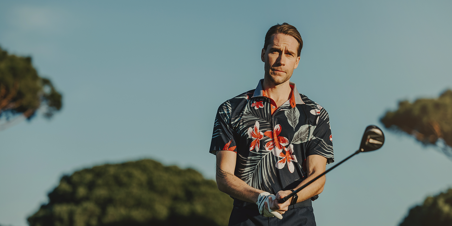 Man in a floral polo shirt holding a golf club outdoors on a golf course with trees in the background.