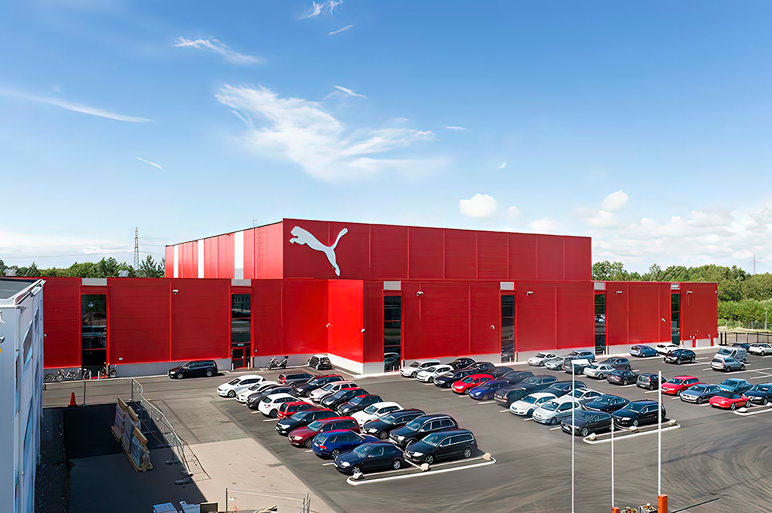 En stor röd byggnad med Puma-logotyp, omgiven av en parkeringsplats fylld med bilar under en klarblå himmel.