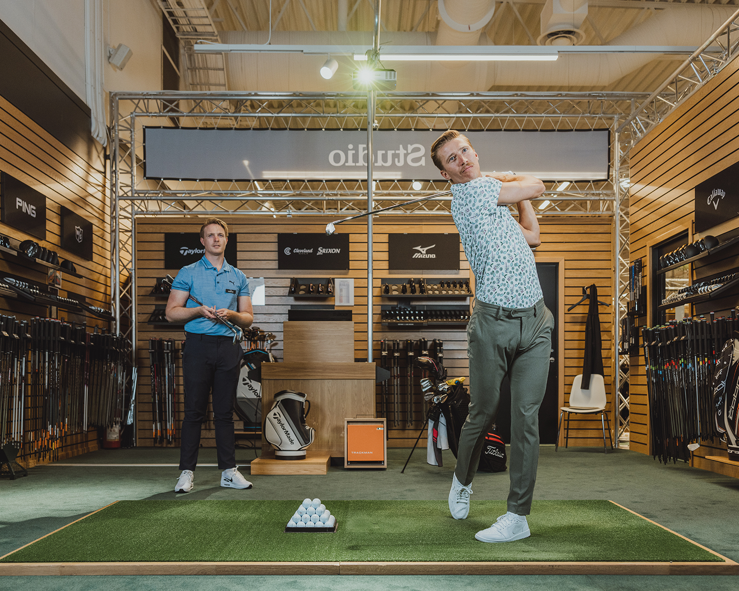 Två män i en golfbutik, en slår en golfboll på en inomhusmatta medan den andra tittar på omgiven av golfklubbor och utrustning.