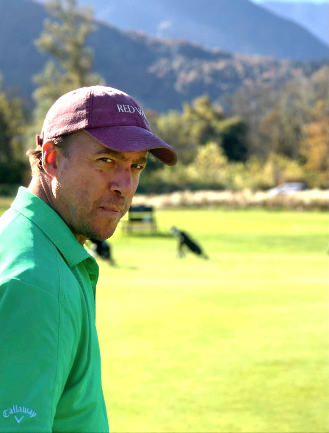 Man i grön pikétröja och vinröd keps med texten "RED" står på en golfbana med berg och träd i bakgrunden.