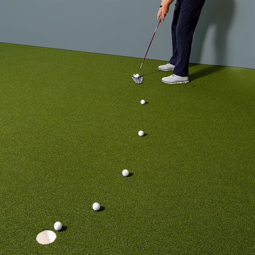 Person håller en golfklubba och tränar puttning på konstgräs med flera golfbollar i rad mot ett hål.