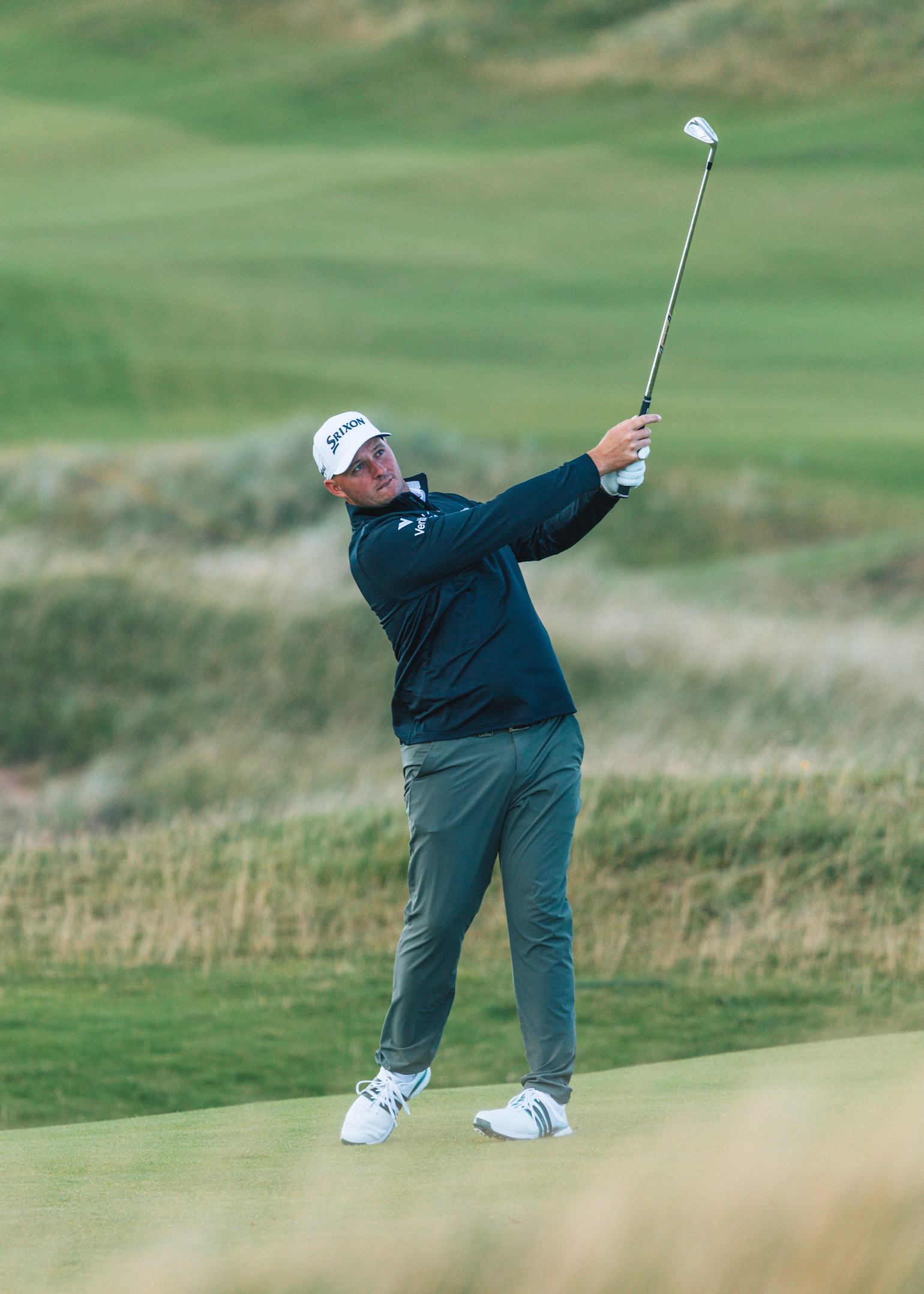 Golfspelare svingar en klubba på en grön golfbana, klädd i mörk jacka, keps och sportskor.