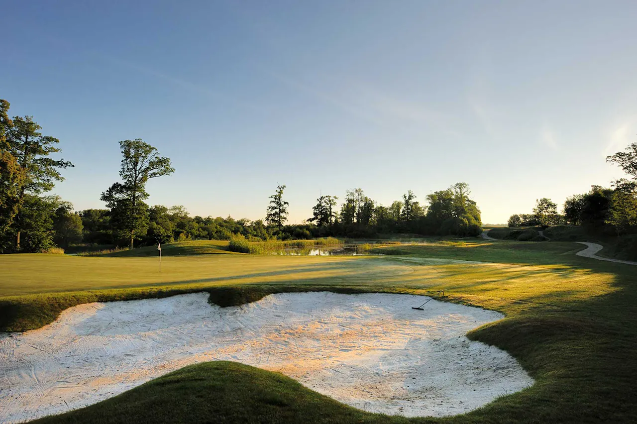 Sandbunker och green på en golfbana med flaggstång, omgiven av gräs, träd och en liten damm i bakgrunden vid solnedgång.