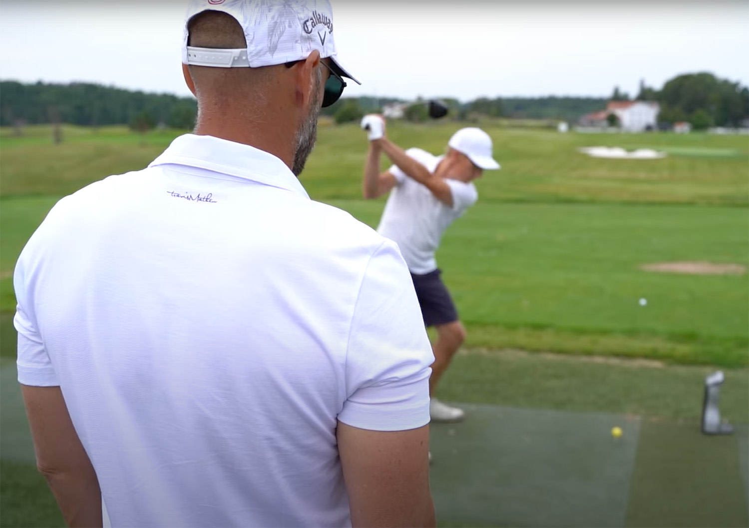 Två män i vita kepsar och pikétröjor tränar golf på driving range, en slår medan den andra tittar på.