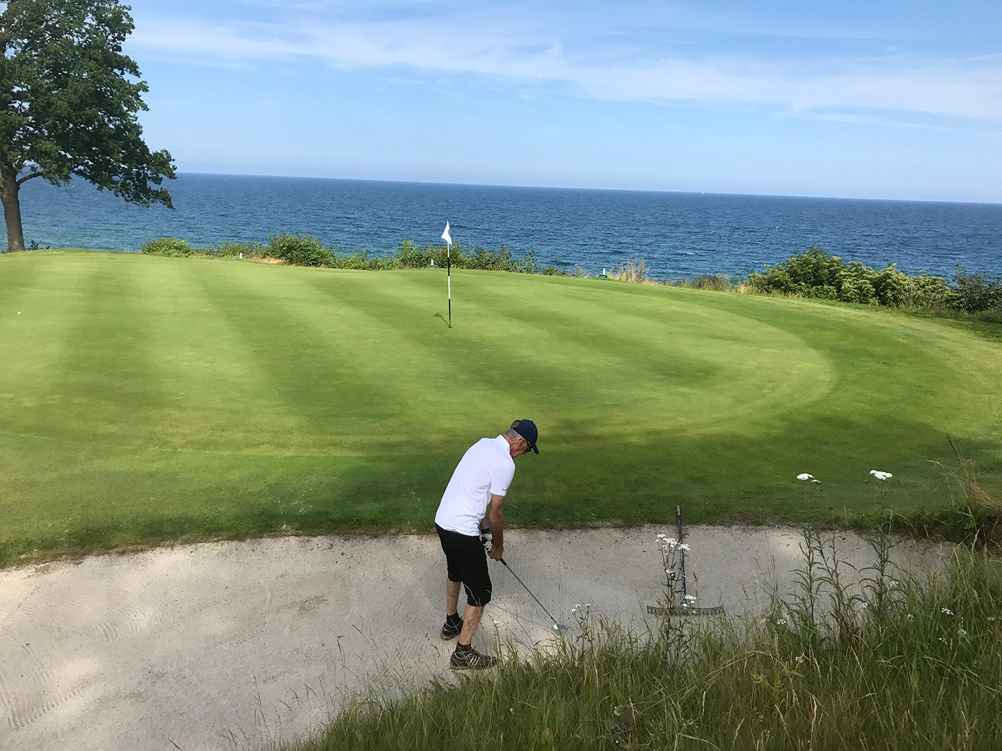 Golfspelare slår ett slag från en sandbunker mot greenen med havet och blå himmel i bakgrunden.