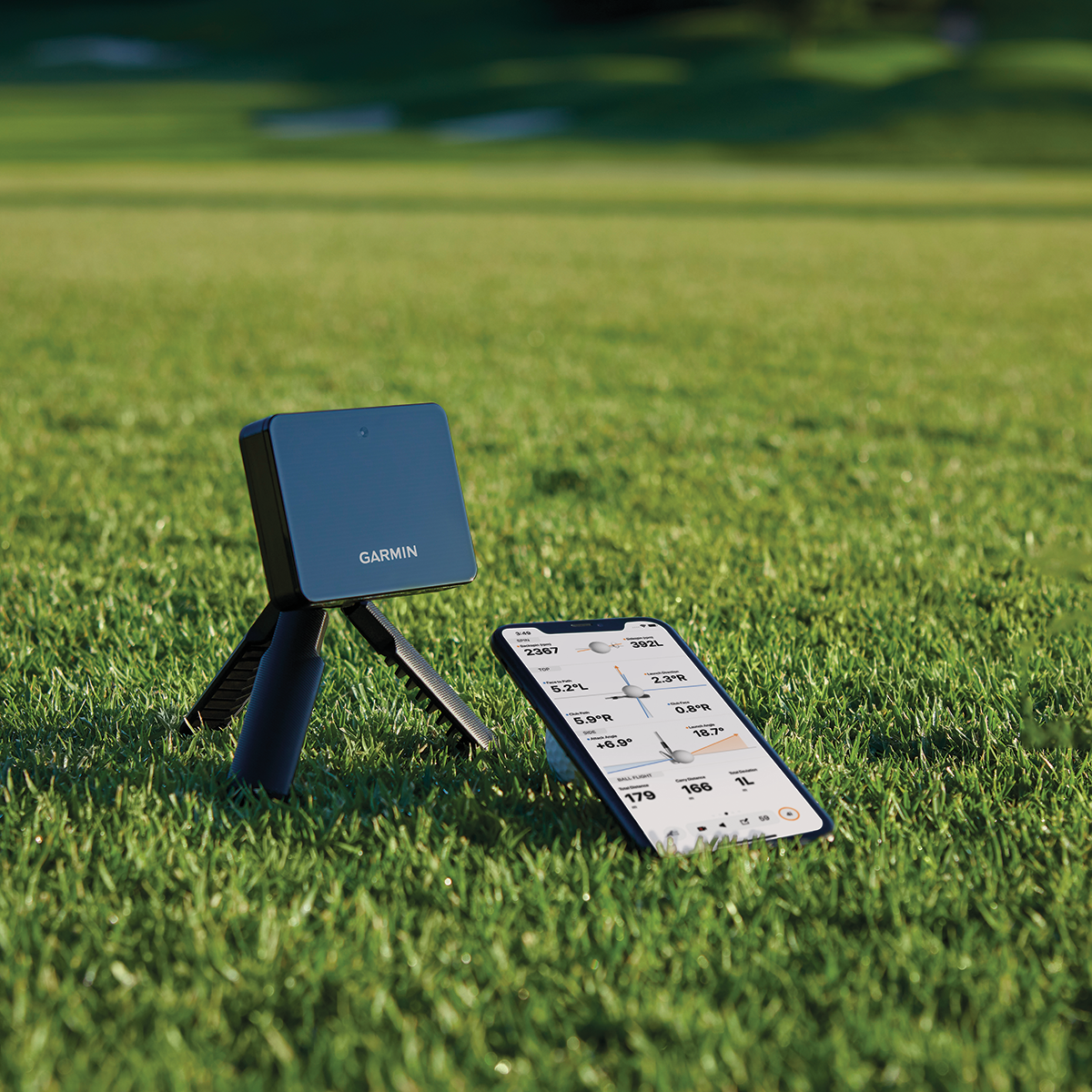 En Garmin-enhet på stativ står på en golfbana bredvid en smartphone som visar golfdata på skärmen.