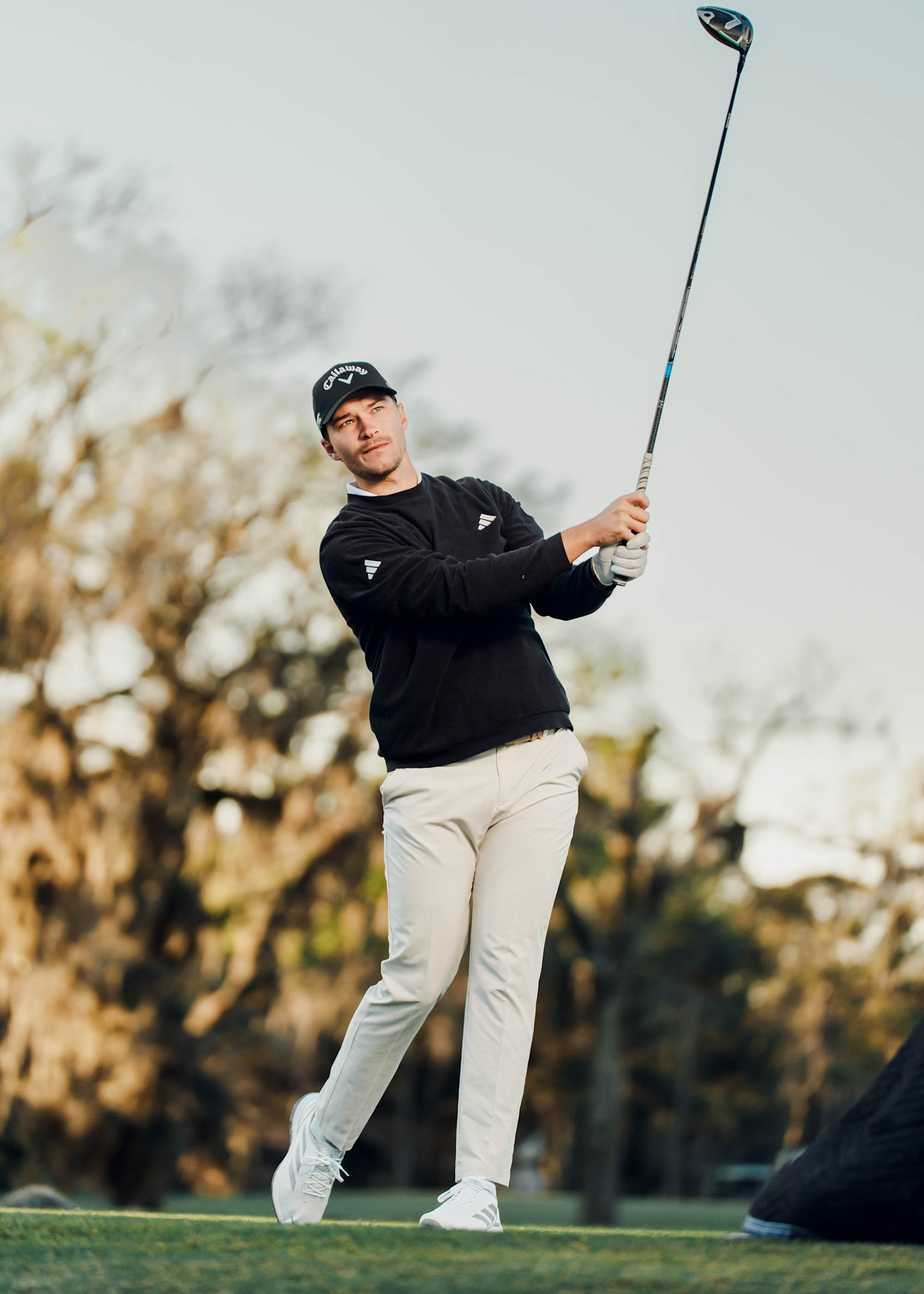 Man som svingar en golfklubba på en golfbana, klädd i svart tröja, ljusa byxor och keps, med träd i bakgrunden.