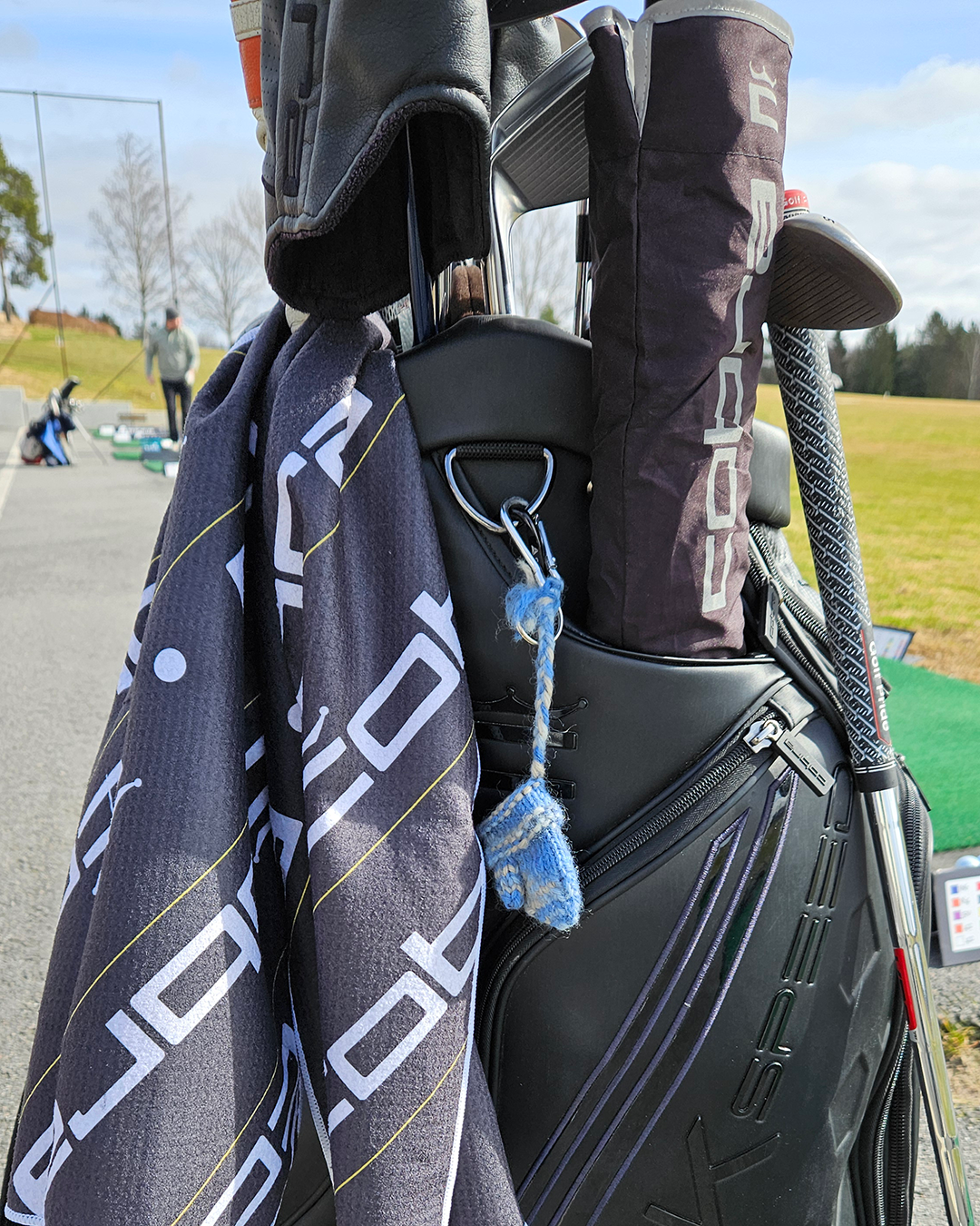 Golfbag med klubbor, handduk med vit text, och klubbskydd på driving range med gräs och människor i bakgrunden.