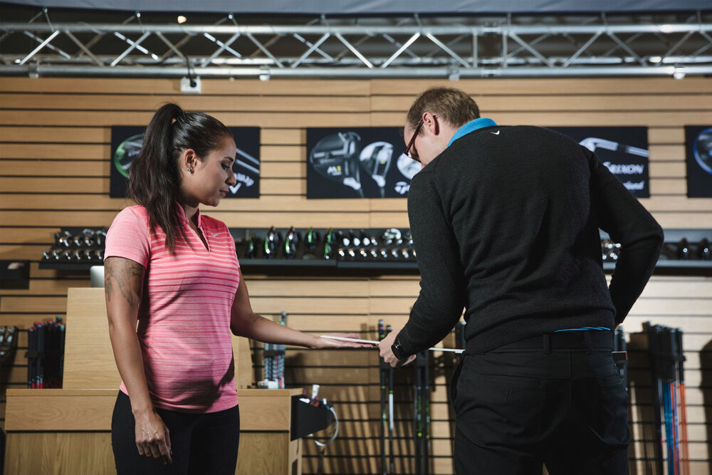 En kvinna i rosa tröja får hjälp av en man med att mäta sin hand i en golfbutik med klubbor i bakgrunden.