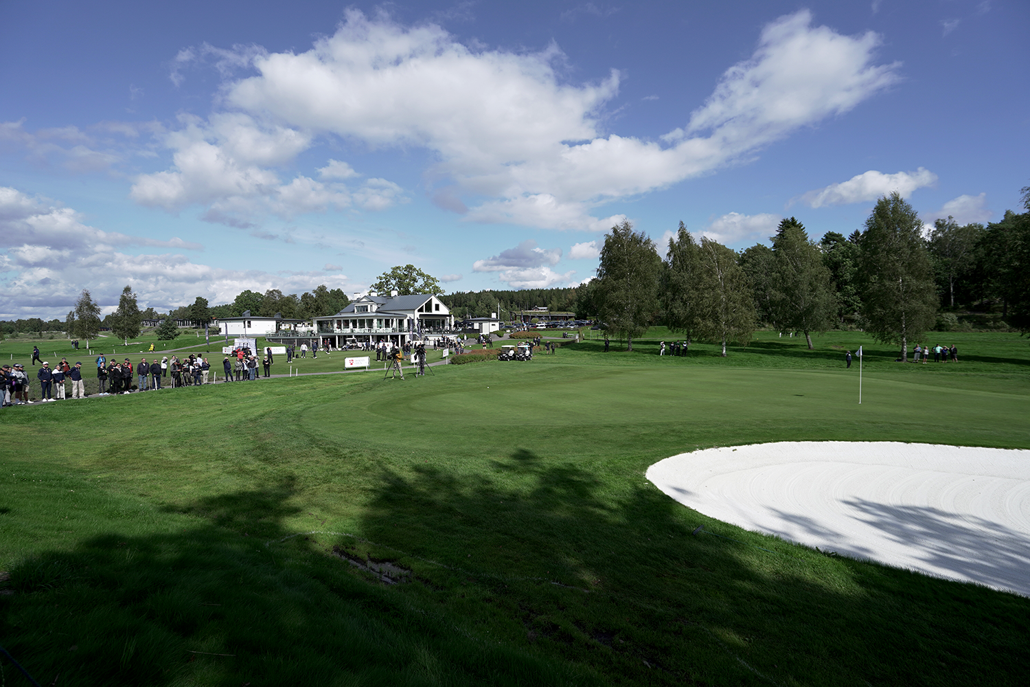 Publik samlas vid en golfbana nära klubbhuset, med sandbunker och green i förgrunden under en blå himmel.