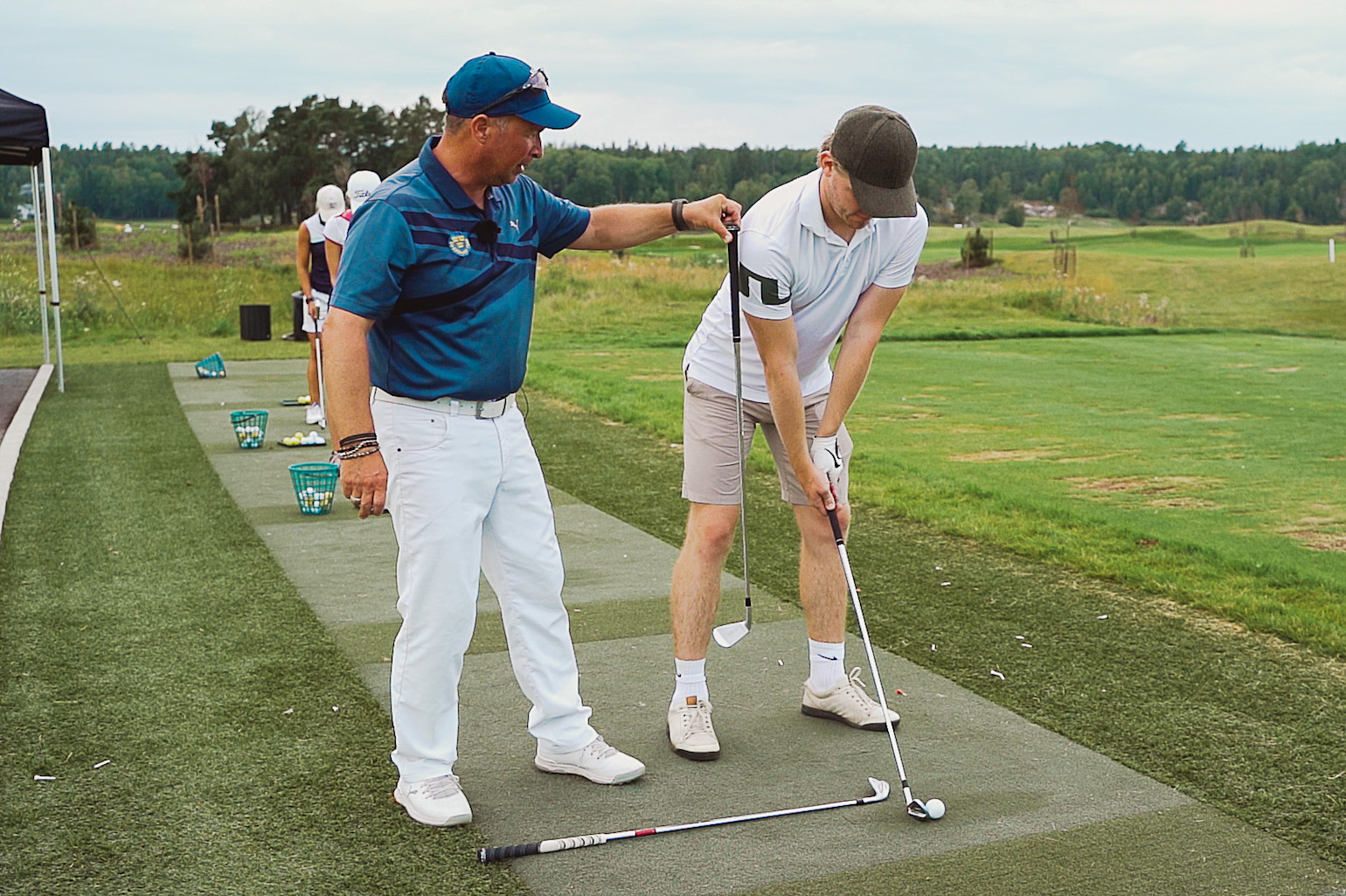 Golftränare instruerar en elev i rätt svingteknik på en driving range, med golfbollar och korgar i bakgrunden.